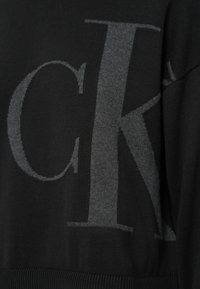 Melns adīts džemperis ar lielu pelēku logotipa rakstu, teksturētu audumu, ribveida apakšu un manžetēm. Tuvplāns uz dizaina detaļām.