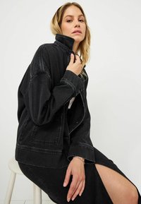 Svart denimjacka med hög krage, oversize passform och framfickdetaljer. Bärs över en svart kjol med en sidssprund. Slät textur.