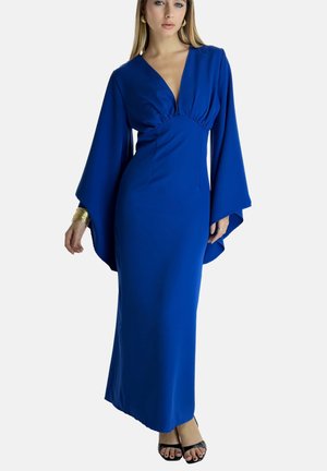 Lange blauwe jurk met een diepe v-hals, een strakke taille en brede mouwen. De soepele stof valt tot de enkels en is gecombineerd met zwarte hakken.