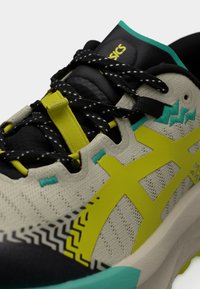 Κοντινή λήψη ενός δρομικού παπουτσιού σε μπεζ και μαύρο χρώμα με κίτρινες πινελιές, μαύρα κορδόνια με λευκές βούλες και τιρκουάζ λεπτομέρειες στη σόλα και το πάνω μέρος.