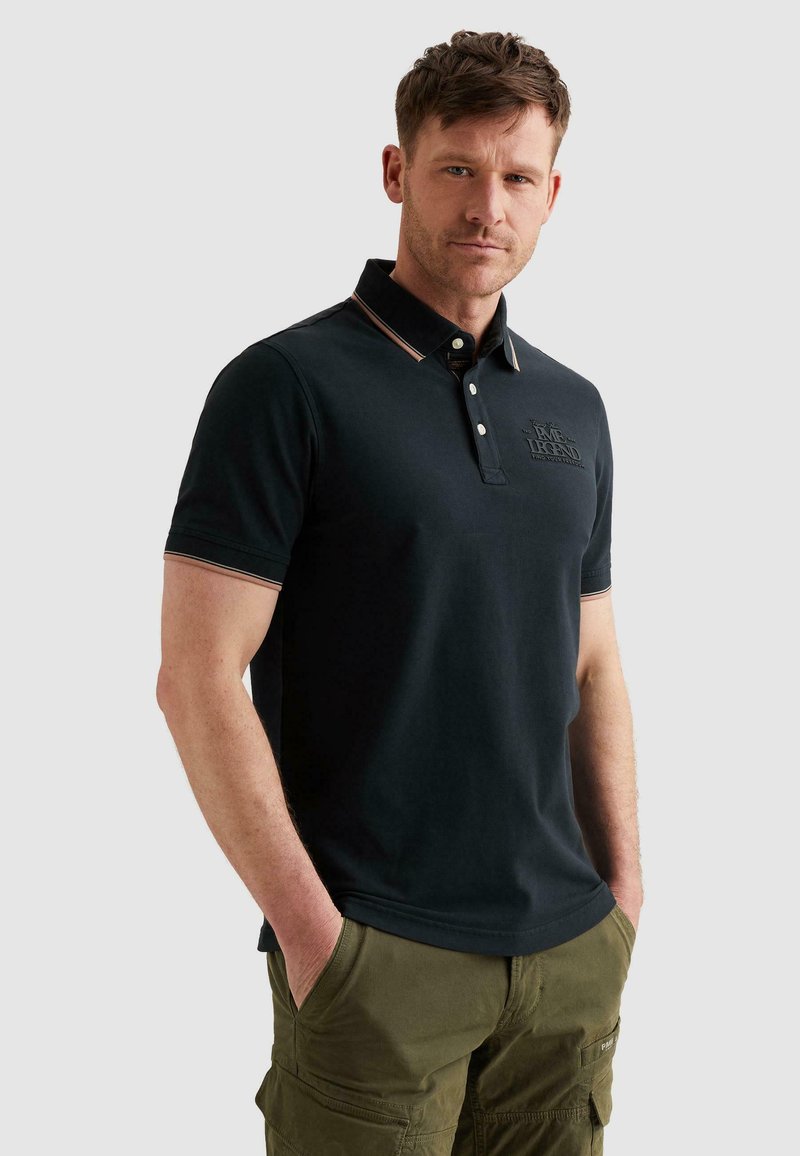 Homme portant un polo noir à manches courtes avec un logo brodé et un pantalon cargo vert olive, debout les mains dans les poches.