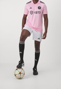 Vaaleanpunainen jalkapallon pelipaita mustilla viivoilla, valkoiset shortsit, mustat sukat valkoisilla viivoilla ja mustat nappulakengät monivärisen jalkapallon päällä.