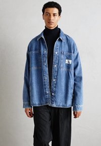 Jachetă din denim de culoare albastră deschisă, cu două buzunare frontale și o patch cu logo. Are un croi lejer și închidere cu nasturi. Purtați-o peste un pulover cu guler înalt de culoare neagră.