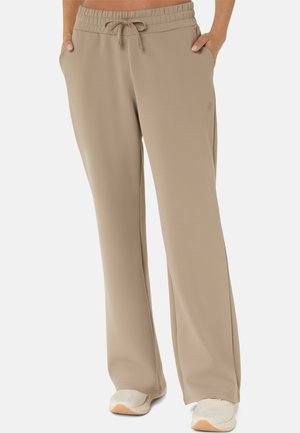 Beige vidde bukser med elastisk talje og snøre, model med hænder i lommerne, iført hvide sneakers.