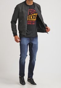 Chaqueta bomber negra con puños acanalados, camiseta negra con texto amarillo y rojo, jeans azules y zapatillas negras con suelas blancas.