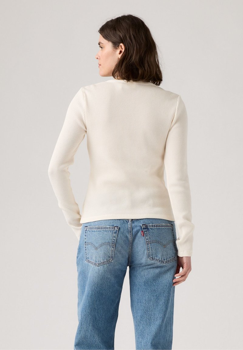 Camisa de manga larga de canalé crema, diseño ajustado, cuello redondo, textura suave; combinada con jeans de tiro alto azul claro que cuentan con bolsillos traseros.