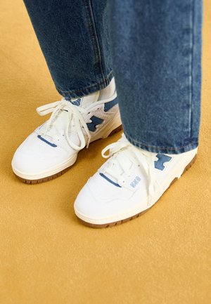 Witte sneakers met blauwe accenten en bruine zolen, gedragen met blauwe spijkerbroeken op een beige tapijtvloer.