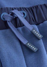 Ceinture de pantalon de survêtement en maille bleue avec des cordons tissés passant dans des œillets, marquée "BAKER" sur les embouts en plastique.