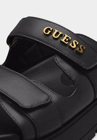 Μαύρο δερμάτινο σανδάλι με διπλά λουριά και χρυσά γράμματα «GUESS» στο πάνω λουρί, που δείχνει μαξιλαρωμένο πάτο και χοντρή σόλα.
