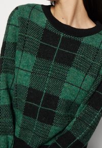 Pull tricoté avec un motif à carreaux verts et noirs. Il présente un col rond côtelé et une finition texturée, mettant en valeur des tissages de tissu variés.