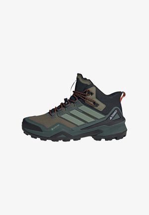 Shoe de caminhada de cano alto com parte superior em verde escuro e preto, painéis de malha texturizados, riscas cinzas, detalhes laranja e sola preta robusta.