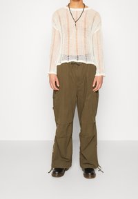 Person klädd i en genomskinlig vit stickad tröja, olivgröna cargobyxor med sidofickor, svarta stövlar och ett svart pärlhalssmycke med en hänge.