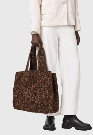 Άτομο με λευκή ενδυμασία και μαύρες μπότες κρατά μια μεγάλη καφέ και μαύρη τσάντα ώμου με τύπωμα λεοπάρδαλης και φαρδιές λαβές.