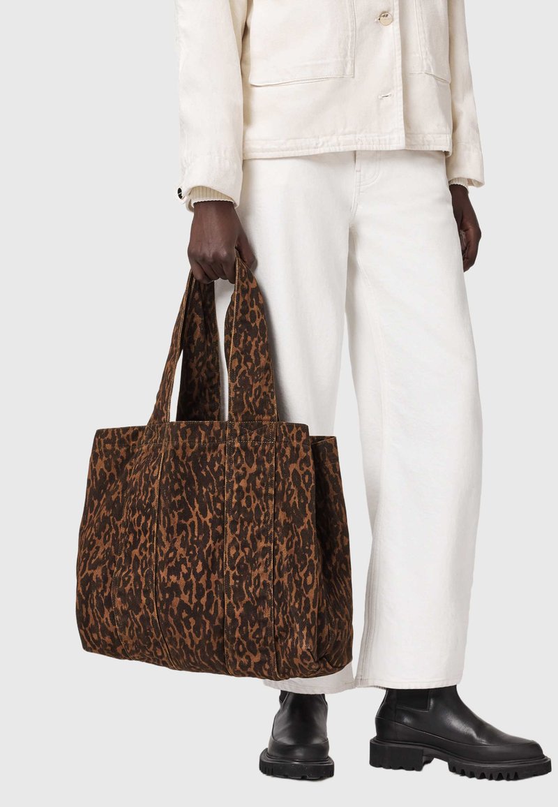 Άτομο με λευκή ενδυμασία και μαύρες μπότες κρατά μια μεγάλη καφέ και μαύρη τσάντα ώμου με τύπωμα λεοπάρδαλης και φαρδιές λαβές.