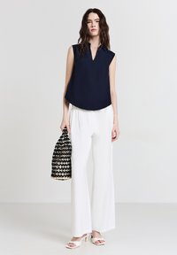 Donna con capelli lunghi e ondulati, che indossa un top senza maniche blu navy, pantaloni bianchi a gamba larga e tacchi bianchi, mentre tiene in mano una borsa intrecciata nera e beige.