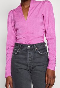 Nainen, jolla on pitkähihainen vaaleanpunainen teksturoitu V-kaula-aukollinen paita, jossa on hienovarainen kuvio, yhdistettynä korkeavyötäröisiin haalistuneisiin mustiin farkkuihin.