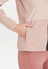 Vaaleanpunainen tuulenpitävä takki, jossa on vetoketjullinen sivutasku ja istuvat hihat. Kankaan pinta on sileä ja hieman kiiltävä.