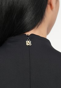 Czarny top z tkaniny o wysokim dekolcie i zamku błyskawicznym z tyłu, z złotym napisem "HUGO" jako elementem wykończenia.