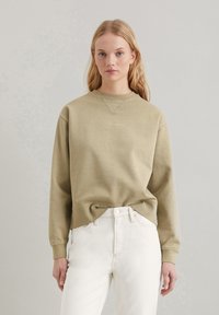 Olivgrüner Pullover mit Rundhalsausschnitt und gerippten Manschetten, kombiniert mit weißen, hochgeschnittenen Jeans. Einfaches Design mit minimalem Branding in Gold.