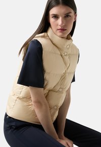 Gepolsterte Weste in hellem Beige mit hohem Kragen, vorderen Druckknöpfen und einem gesteppten Design. Getragen über einem navyfarbigen Kurzarmoberteil.