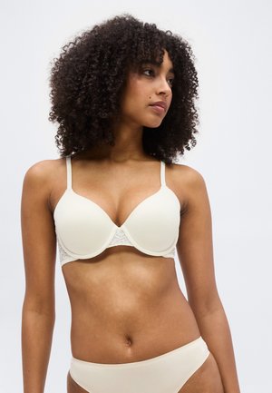 Donna con capelli ricci che indossa un reggiseno color crema con dettagli in pizzo e mutande abbinate, che guarda verso destra su uno sfondo semplice.