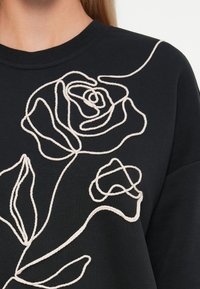 Primer plano de una prenda negra con bordado de cuerda beige que forma un diseño abstracto de rosa y hojas en el pecho.