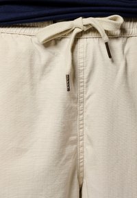 Shorts beiges avec un motif à carreaux texturé, dotés d'une taille à cordon avec des liens en tissu et des embouts métalliques, associés à une chemise bleu marine.