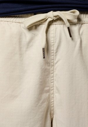 Shorts beiges avec un motif à carreaux texturé, dotés d'une taille à cordon avec des liens en tissu et des embouts métalliques, associés à une chemise bleu marine.