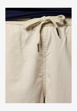 Shorts beiges avec un motif à carreaux texturé, dotés d'une taille à cordon avec des liens en tissu et des embouts métalliques, associés à une chemise bleu marine.