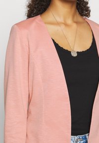 Světle růžový blazer s hladkým povrchem, otevřeným předním designem a dlouhými rukávy. Nosený přes černý žebrovaný top s krajkovým lemem kolem krku.