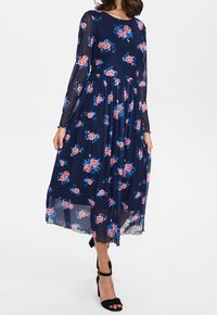Vestido floral marino con mangas largas de tul, decorado con rosas rosas y acentos azules, falda fluida y dobladillo con ondas. Tacones altos negros.