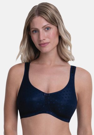 Vrouw met schouderlang blond haar die een donkerblauwe beha draagt met subtiele polkadots en kleine decoratieve knoopjes op de bandjes en in het midden.