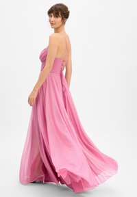 Frau in einem glitzernden rosa bodenlangen Kleid mit überkreuzten Rückenträgern, schaut über ihre Schulter vor einem schlichten weißen Hintergrund.