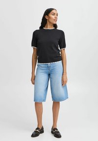 Sort kortærmet top med hvid bølget trim, parret med lyseblå denim bermudashorts og sorte loafers med guld accenter.