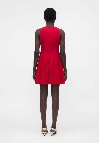 Γυναίκα με κοντά σγουρά μαλλιά, ντυμένη με ένα αμάνικο κόκκινο φόρεμα μέχρι το γόνατο και λευκά πέδιλα με ψηλά τακούνια, στέκεται γυρισμένη πλάτη σε μονόχρωμο φόντο.