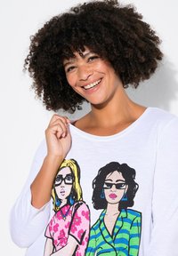 Maglietta bianca a maniche lunghe con illustrazioni grafiche colorate di due donne; una con un top floreale e l'altra con un design a righe.