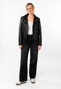 Giacca da motociclista in pelle nera con cerniere argento, abbinata a jeans in denim neri a vita alta e sneakers bianche con righe nere.