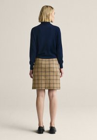 Nainen tummansininen pitkähihainen paita, beige ruudullinen hame ja mustat kengät, seisoo selkä kameraan päin yksinkertaisen beigen taustan edessä.