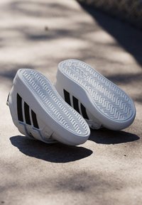 Bijele sportske tenisice s crnim prugama, s teksturiranim gumenim potplatom i elegantnim dizajnom, prikazane s donje i bočne strane.