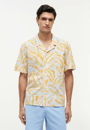 Mand iført kortærmet skjorte med gult palmebladsmønster og lyseblå shorts, stående foran en ensfarvet baggrund.