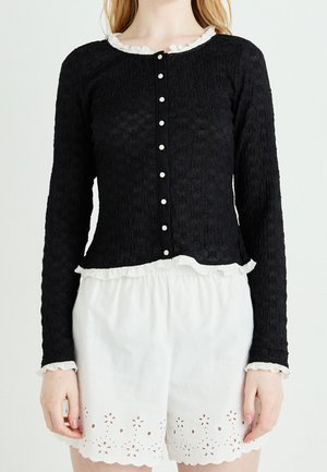 Cardigan noir texturé à manches longues avec boutons blancs et bords volantés, associé à un short blanc à ourlet festonné avec découpes florales en broderie anglaise.