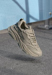 Beige sneakers featuring a smooth upper with mesh accents, thick sole with distinct traction pattern, and laces for a secure fit.