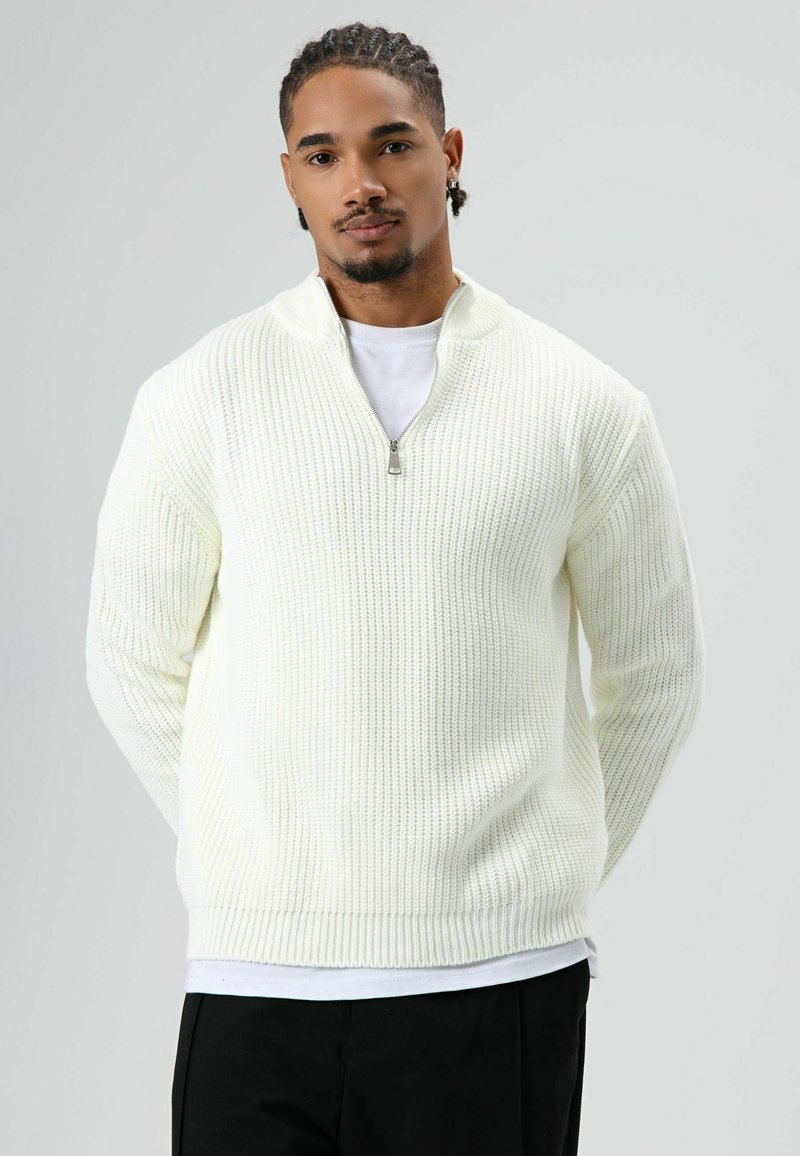 Man with braided hair wears white zip-up knit sweater over white shirt and black pants, standing against plain light background.