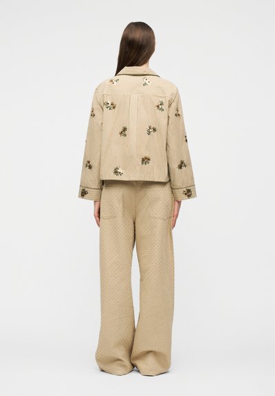 Γυναίκα με μακριά καστανά μαλλιά στέκεται πλάτη, φορώντας μπεζ ριγέ σακάκι με κεντήματα λουλουδιών και μπεζ παντελόνι με πλατιά μπατζάκια και υφή.