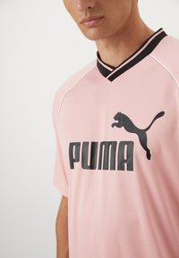 Puma sportinė marškinė šviesiai rožinės spalvos, su juodu logo ir tekstu, juodu V formos apykakle ir trumpomis rankovėmis, pagaminti iš kvėpuojančios medžiagos.