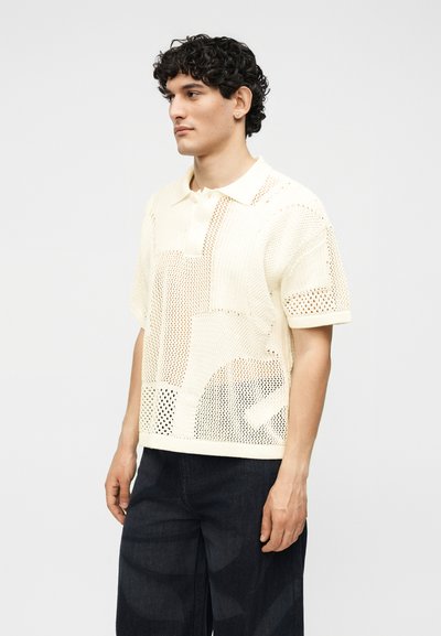 Άνδρας με σγουρά μαλλιά που φοράει ένα κρεμ πλεκτό πόλο με κοντά μανίκια και σκούρο παντελόνι με φαρδιά πόδια, στέκεται μπροστά σε ένα απλό ανοιχτό φόντο.