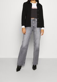 Chaqueta negra de botones sobre un top corto gris, combinada con unos pantalones anchos de talle alto en gris claro y botas de tacón negras. Textura suave.