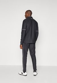 Survêtement de sport noir avec des accents blancs. Comprend une veste zippée et un pantalon assorti avec des bandes latérales. Le tissu semble lisse et flexible.