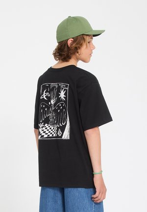 Adolescent portant une casquette verte, un T-shirt noir avec un graphisme blanc d'une figure ailée et du texte, et un jean bleu, se tenant de profil sur un fond blanc.
