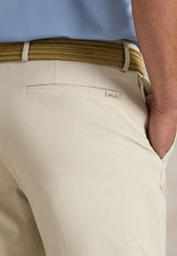 Pantalones beige confeccionados con tela suave que presentan un bolsillo trasero y una etiqueta de marca. Se lleva un cinturón texturizado y tejido de tono marrón claro.